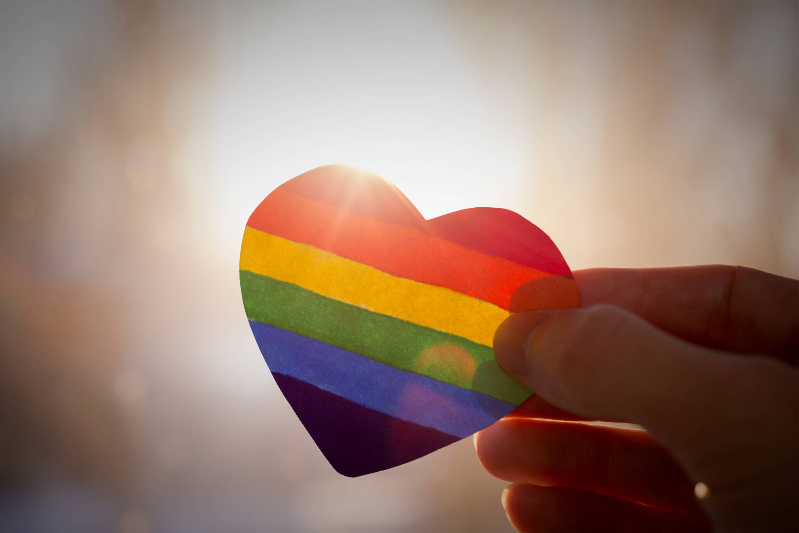 A rainbow shaped heart being held by a hand up against the sun